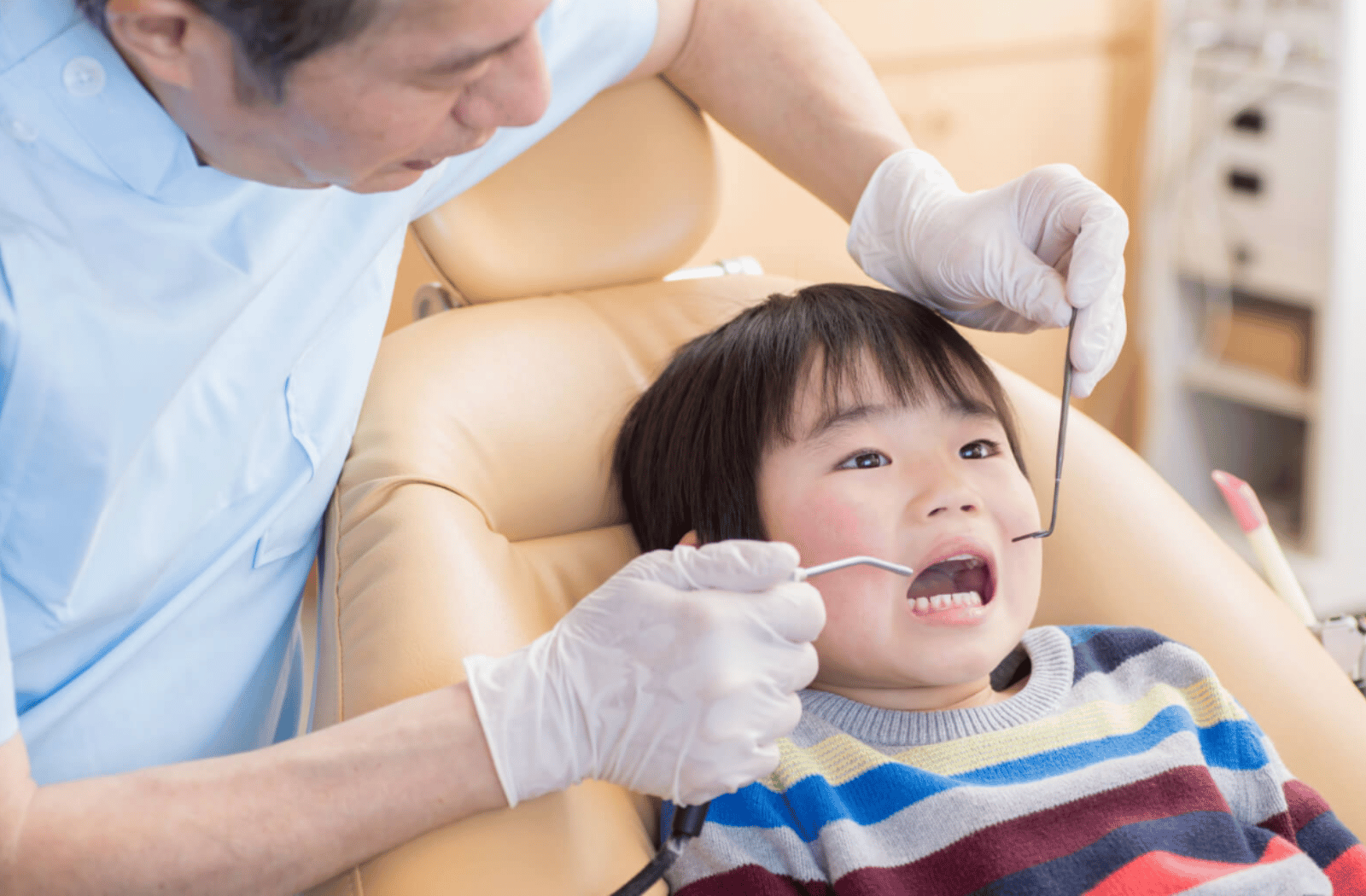 A male dentist is examining a boy's teeth at the dental clinic. The boy's mouth is wide open while the dentist is checking if the boy's teeth are okay